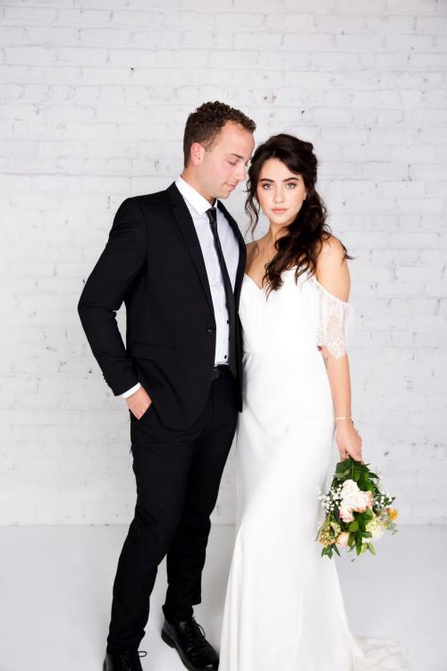 by the Bridal Elite Team Bridal photoshoot of bride wearing a wedding dress with half updo and makeup holding flowers and groom wearing suit and tie