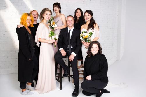 Behind the scenes of bridal photoshoot stylists and models wearing wedding dress and bridesmaids dresses holding flower bouquet and groom wearing suit and tie