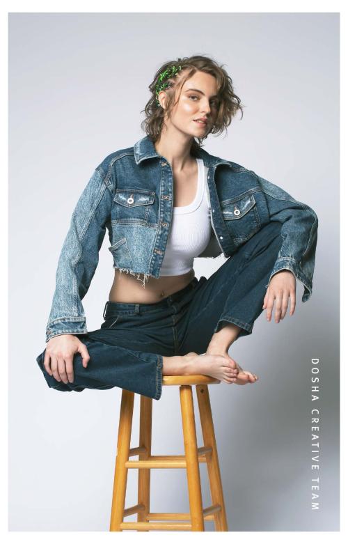 Braids x Denim photoshoot by Dosha Creative Team with a young woman sitting on a wooden stool with her feet up wearing jean pants and a cropped denim jacket. Woman has short blonde hair with a side braid.